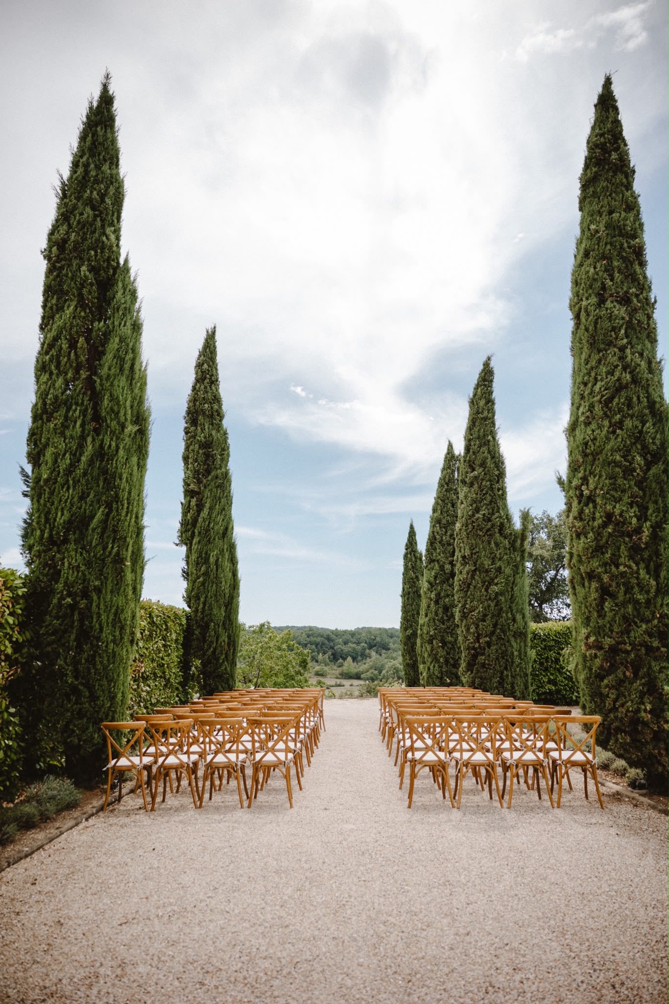 Rows of wooden chairs along a gravel aisle lined with cypress trees